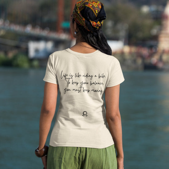 A person stands by a river wearing the eco-friendly "Pedal through Life" T-shirt featuring the quote, "Life is like riding a bike. To keep your balance, you must keep moving." The individual has long hair partially covered by a patterned scarf and wears green pants. A bridge and blurred trees are visible in the background.