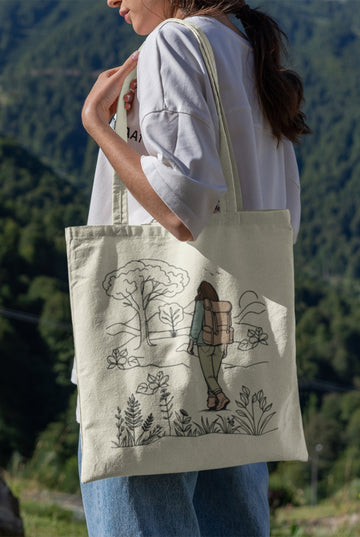 An adventurous woman stands outside, clutching her eco-friendly The Adventurous Woman Tote Bag, decorated with a design of a hiker exploring a natural setting surrounded by trees and shrubs. She is wearing a white shirt and jeans against the backdrop of forested hills.