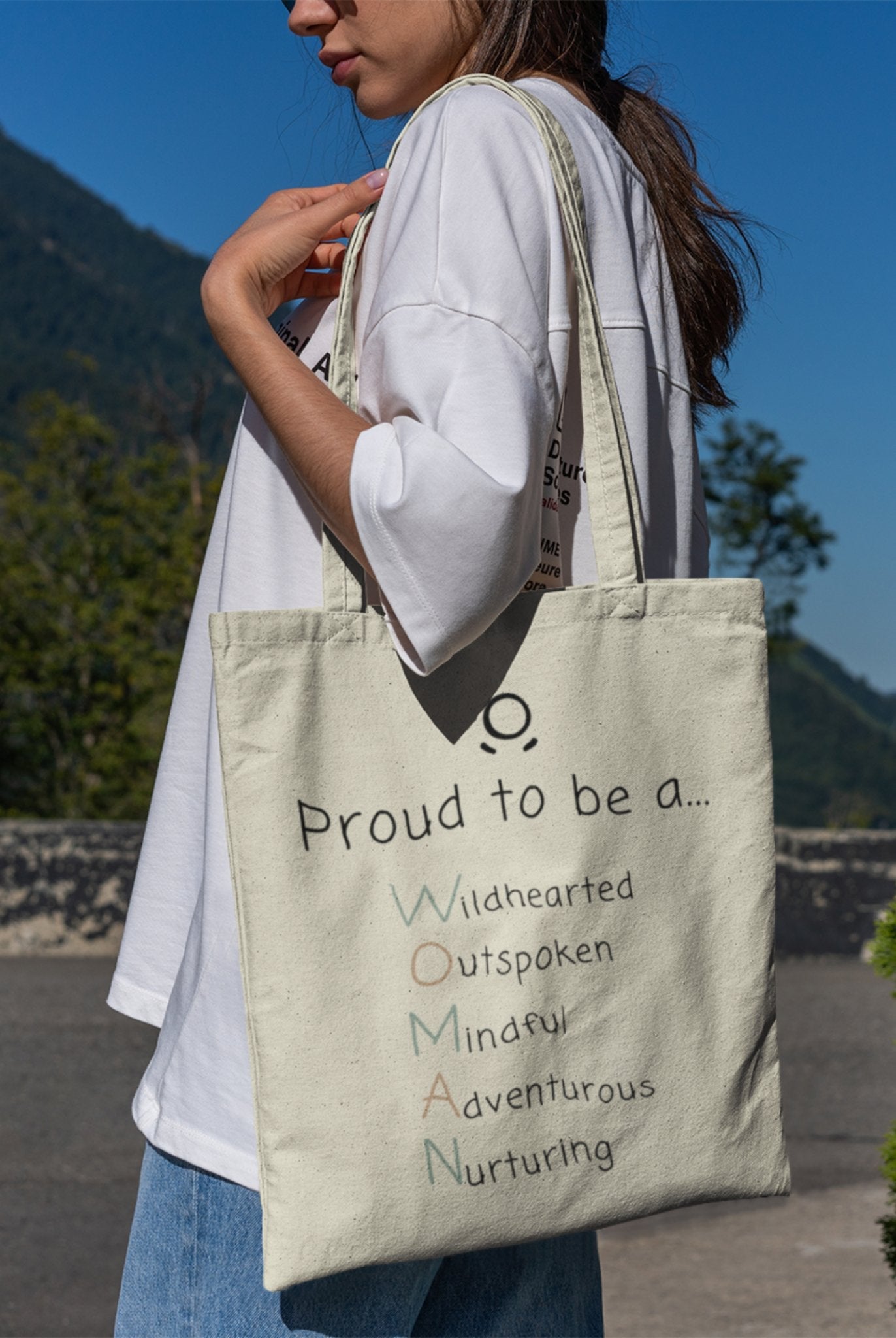 A person holds "The Adventurous Woman Tote Bag," an eco-friendly, cream-colored accessory adorned with the colorful and empowering message: "Proud to be an Adventurous Woman." Each letter symbolizes being Wildhearted, Outspoken, Mindful, Adventurous, and Nurturing. Dressed in a white shirt and jeans, they are outdoors with nature softly fading into the background.