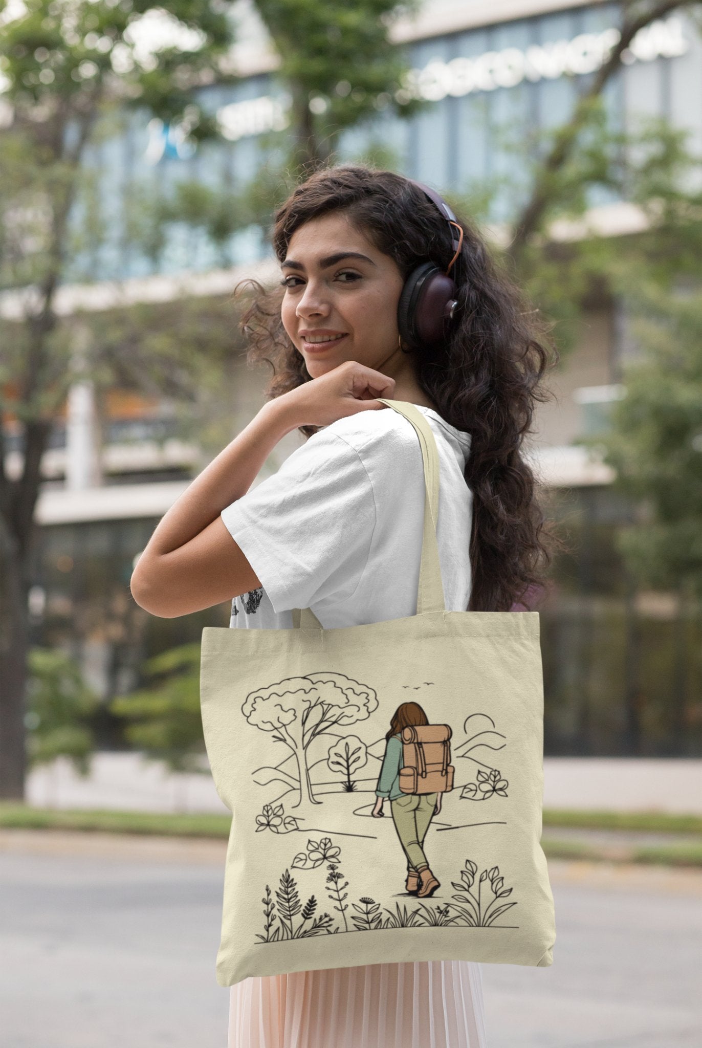 A woman with curly hair, donned in a white t-shirt, smiles as she wears headphones and holds "The Adventurous Woman Tote Bag." This eco-friendly beige tote, ideal for sustainable travel, showcases a design of a person with a backpack exploring nature. She stands on a city sidewalk with trees and blurred buildings in the background.