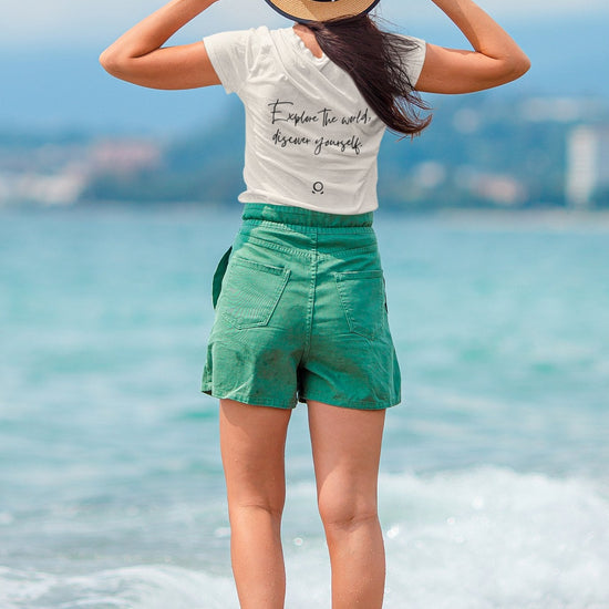 A woman stands on a beach facing the ocean, embodying wanderlust. She wears a straw hat and an eco-conscious Transit Trailblazer Tee with the phrase "Explore the world, Rescue yourself," paired with green shorts. Barefoot in gentle waves, she gazes at the blue sky and distant shoreline.