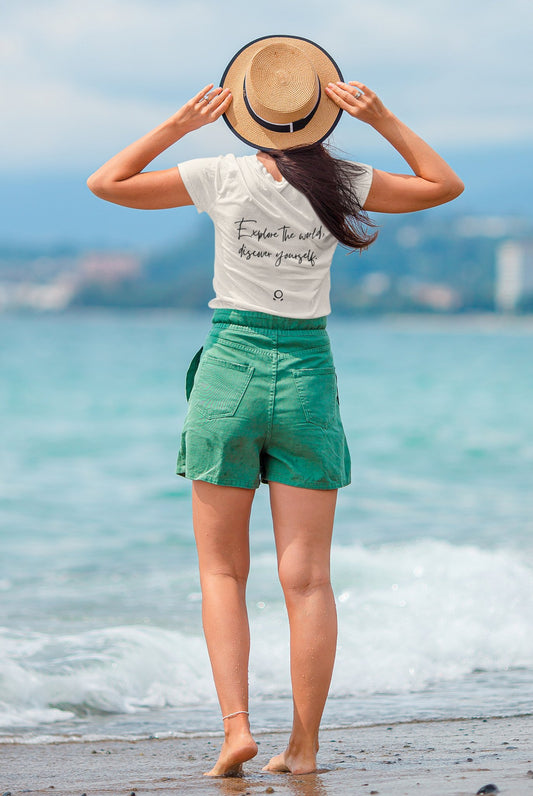 A woman stands on a beach facing the ocean, embodying wanderlust. She wears a straw hat and an eco-conscious Transit Trailblazer Tee with the phrase "Explore the world, Rescue yourself," paired with green shorts. Barefoot in gentle waves, she gazes at the blue sky and distant shoreline.