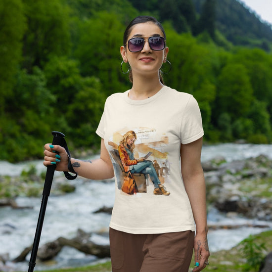 Wearing a Transit Trailblazer Tee and sunglasses, an individual stands by a rocky riverside with a hiking pole in hand, embraced by lush green trees and distant mountains under a clear blue sky. This vibrant and serene setting captures their adventurous spirit and desire for outdoor exploration.