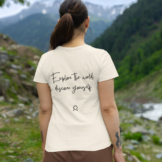 A woman with a ponytail stands outdoors, her eco-conscious Transit Trailblazer Tee adorned with the phrase "Explore the world, discover yourself" as she gazes upon lush green hills and mountains under a cloudy sky. Her spirit of wanderlust perfectly aligns with the breathtaking scenery.