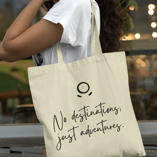 A woman is outdoors, dressed in a white shirt and holding the Adventure Awaits Tote Bag, which features a minimalist logo and eco-friendly design. The background of blurred plants and lights creates an urban, casual atmosphere ideal for sustainable exploration.