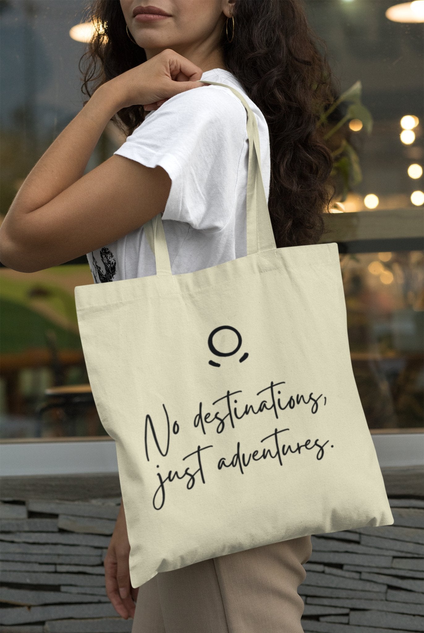 A woman is outdoors, dressed in a white shirt and holding the Adventure Awaits Tote Bag, which features a minimalist logo and eco-friendly design. The background of blurred plants and lights creates an urban, casual atmosphere ideal for sustainable exploration.