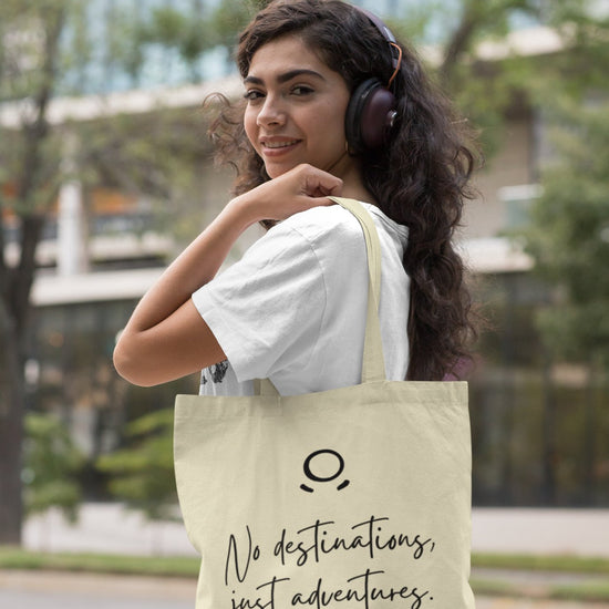 A woman with curly hair smiles as she wears headphones and a white t-shirt, carrying the Adventure Awaits Tote Bag adorned with the phrase "No destinations, just adventures." The backdrop features a blurred outdoor city scene with trees and a building, ideal for sustainable exploration.