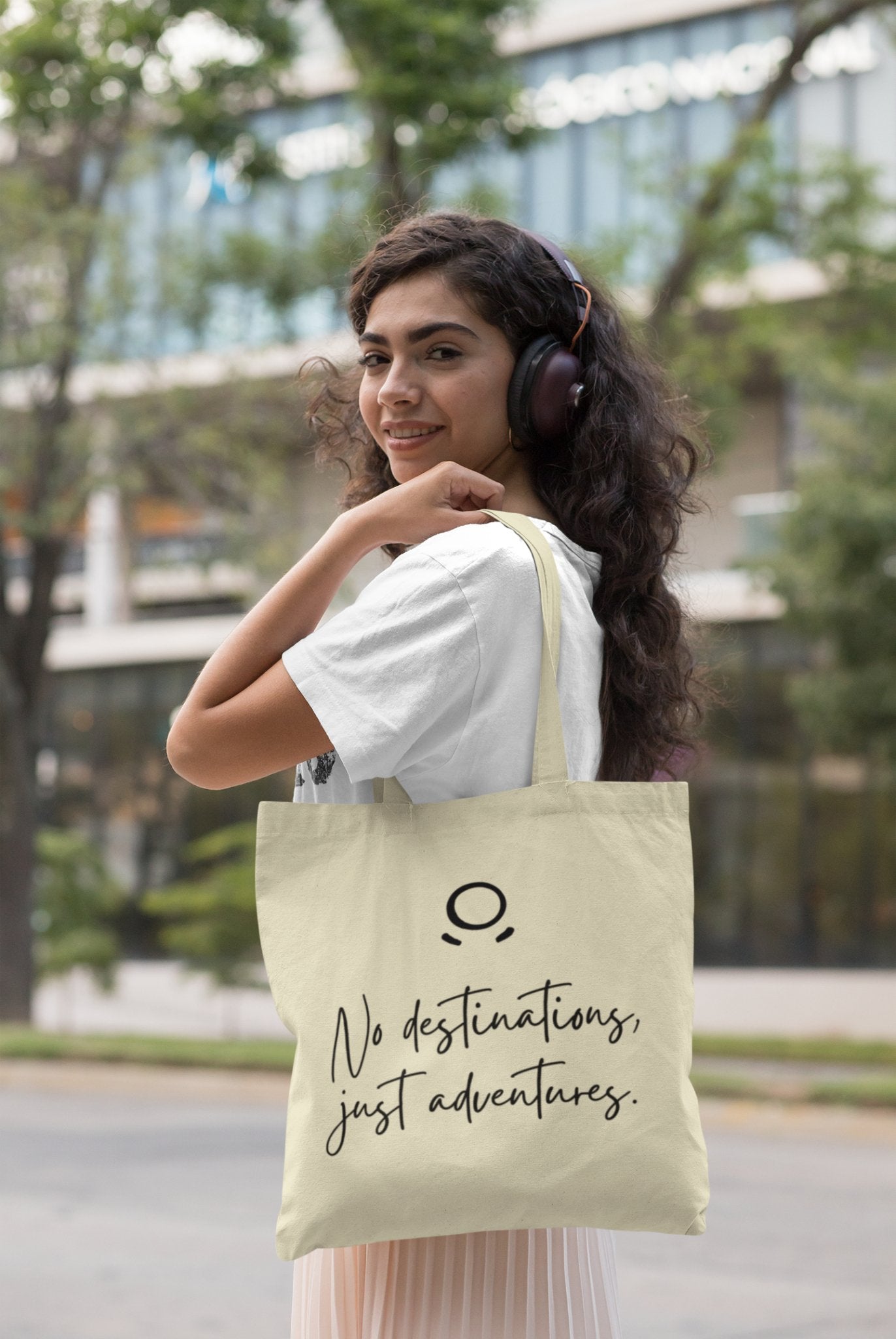 A woman with curly hair smiles as she wears headphones and a white t-shirt, carrying the Adventure Awaits Tote Bag adorned with the phrase "No destinations, just adventures." The backdrop features a blurred outdoor city scene with trees and a building, ideal for sustainable exploration.