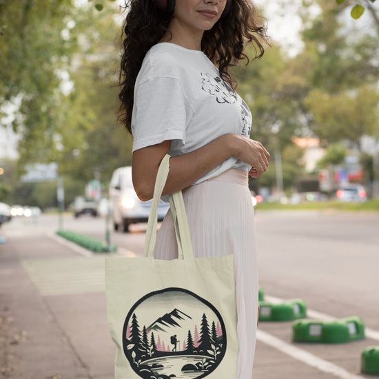 A woman with long hair and glasses, donning headphones, stands on a sidewalk holding the Adventure Awaits Tote Bag, which showcases a mountain and forest design. She's dressed in a white t-shirt and light pink skirt, with trees and cars visible in the background, capturing the essence of sustainable exploration.