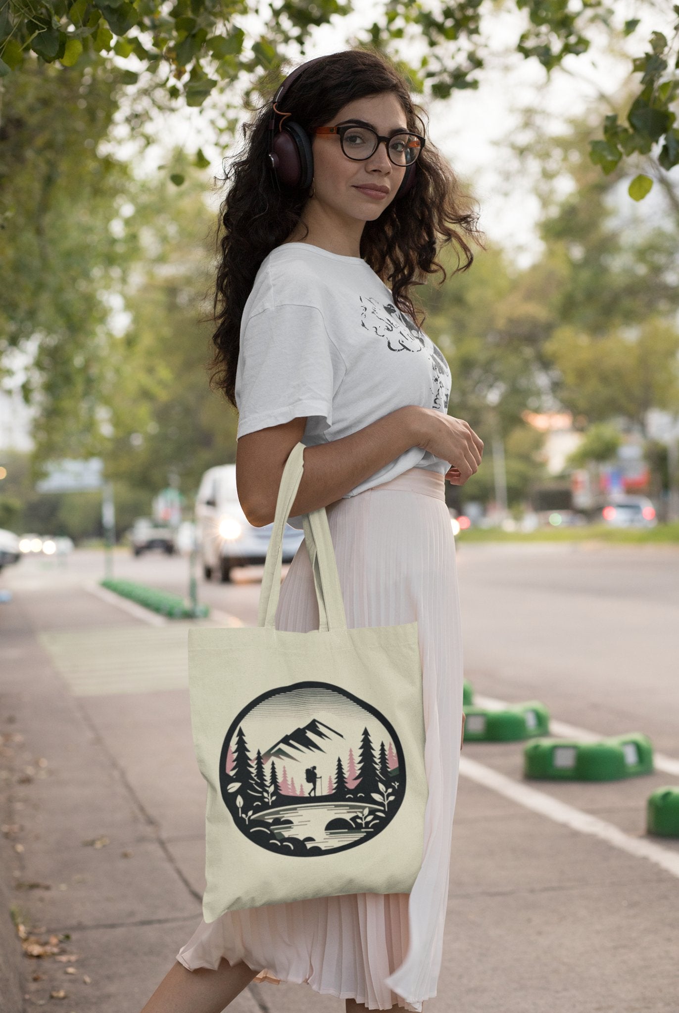 A woman with long hair and glasses, donning headphones, stands on a sidewalk holding the Adventure Awaits Tote Bag, which showcases a mountain and forest design. She's dressed in a white t-shirt and light pink skirt, with trees and cars visible in the background, capturing the essence of sustainable exploration.