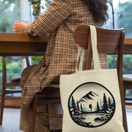 A person in a plaid coat sits at a wooden table with their back to the camera. The Adventure Awaits Tote Bag, featuring an eco-friendly design with a mountain scene graphic—complete with trees, a hiker, and a bridge—hangs from the chair. Large windows reveal greenery outside, hinting that adventure awaits just beyond the glass panes.