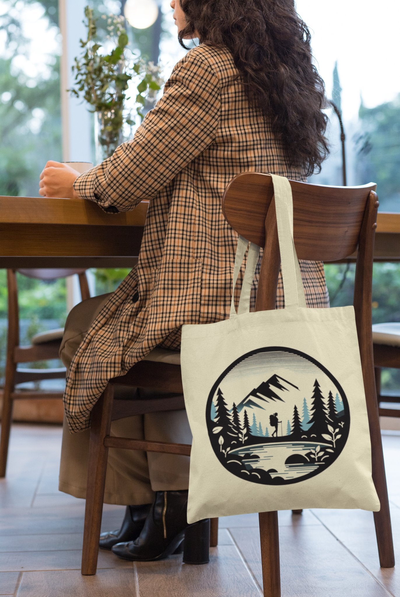 A person in a plaid coat sits at a wooden table with their back to the camera. The Adventure Awaits Tote Bag, featuring an eco-friendly design with a mountain scene graphic—complete with trees, a hiker, and a bridge—hangs from the chair. Large windows reveal greenery outside, hinting that adventure awaits just beyond the glass panes.