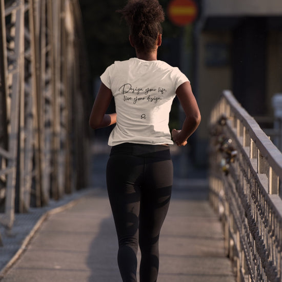 From behind, a person is seen jogging on a bridge, embodying their wanderlust in a white "Wanderlust Workspace" tee that says "Design your life like your design," paired with black leggings. The eco-conscious bridge features metal railings and a paved path, beautifully illuminated by the gentle light of early morning or late afternoon.