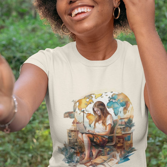 A smiling woman with curly hair takes a selfie outdoors, epitomizing the spirit of a digital nomad. She wears the Wanderlust Workspace Tee, which displays a graphic of a woman using a laptop amid a watercolor-style world map and numerous plants. The vibrant green backdrop complements her wanderlust-inspired setting.