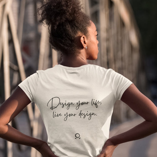 A woman with curly hair styled in a bun stands confidently on a metal bridge, wearing the Wanderlust Workspace Tee, which says "Design your life, live your design." Channeling the essence of digital nomads and an eco-friendly lifestyle, she gazes away from the camera with her hands on her hips.